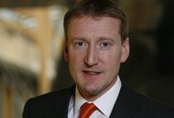 The image shows Tavish Scott facing at the camera and smiling in front of a brown background