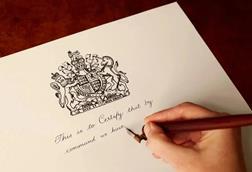 A hand holding a pen and writing on a royal warrant. Text says 'This is to certify that by command we have'
